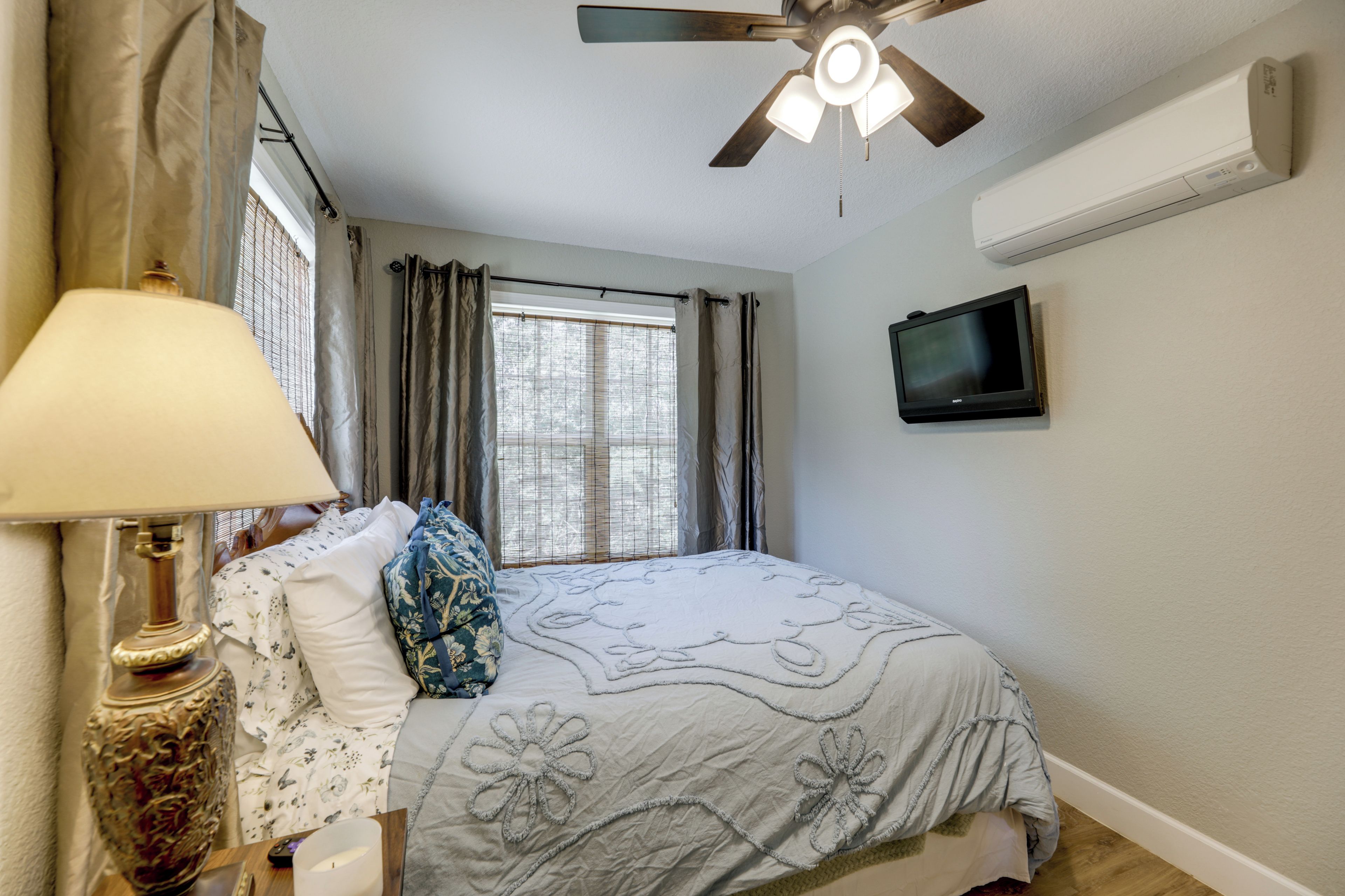 Une chambre à coucher avec un lit, une lampe, un ventilateur de plafond, une fenêtre avec des rideaux et un téléviseur fixé au mur.