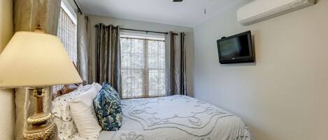 A bedroom with a bed, a lamp, a ceiling fan, a window with curtains, and a television on the wall.