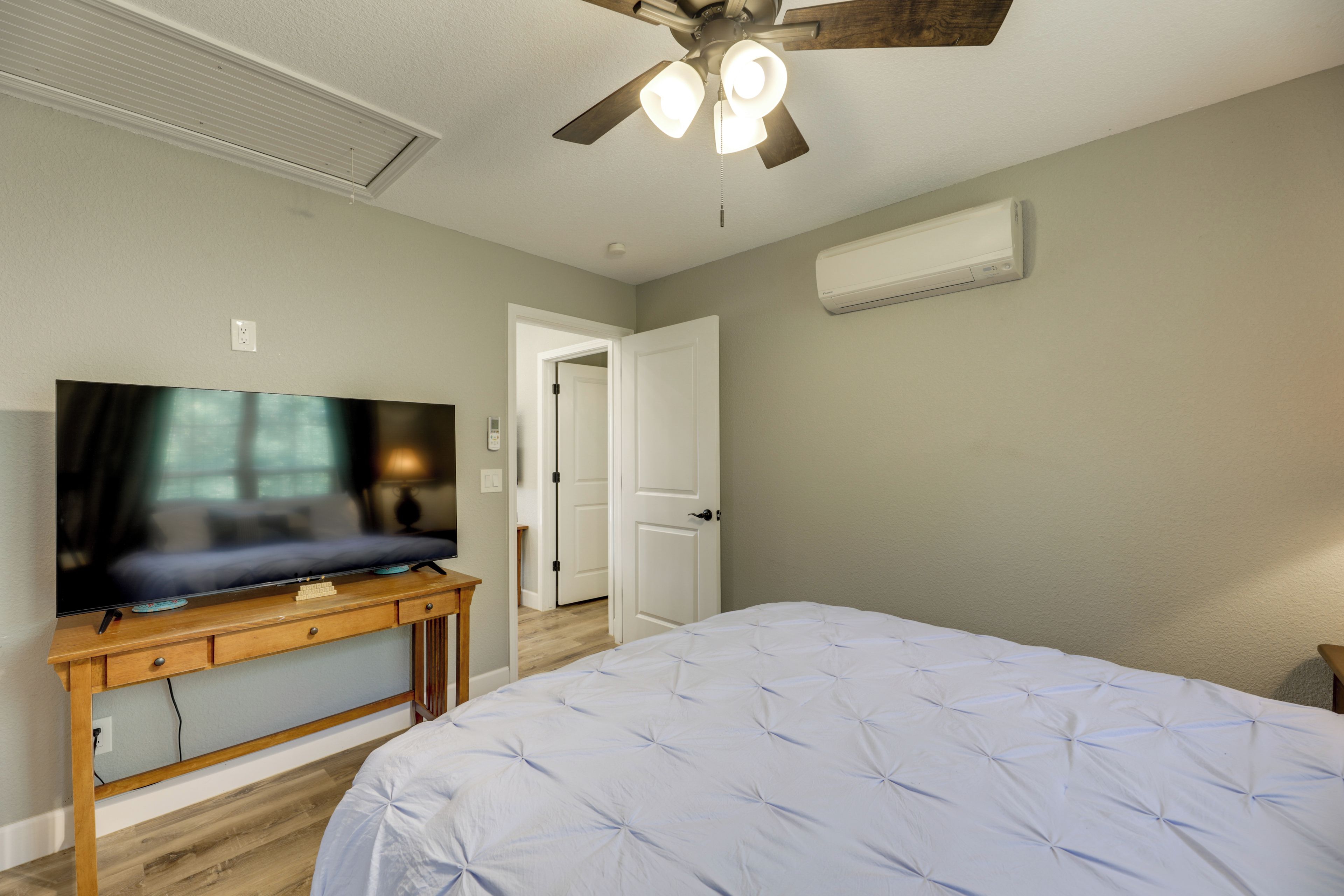 A bedroom with a bed, a TV on a wooden stand, a ceiling fan, and an air conditioning unit.