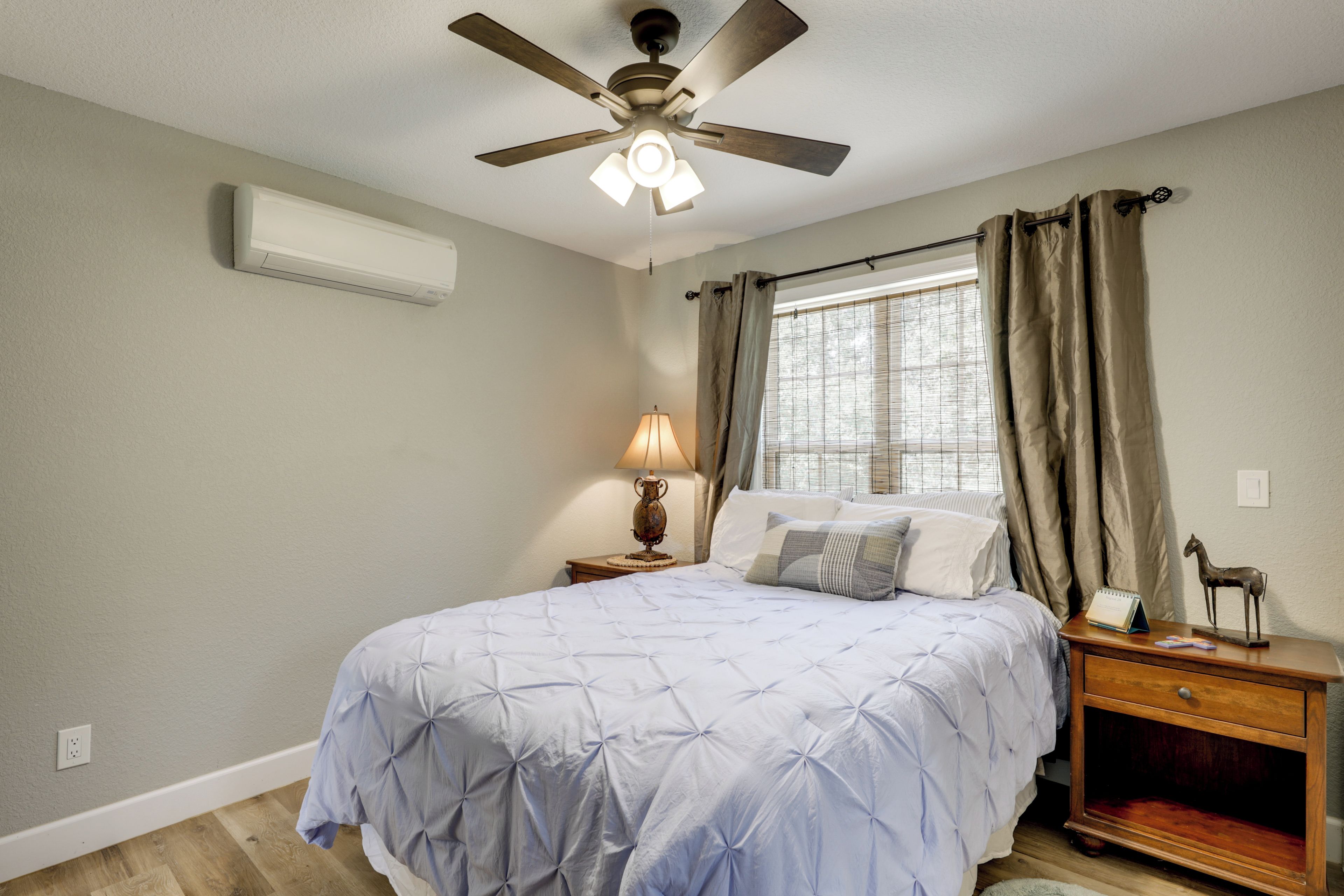 A bedroom with a bed, a nightstand, a lamp, a ceiling fan, a window with curtains, and an air conditioning unit.