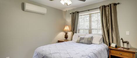 A bedroom with a bed, a nightstand, a lamp, a ceiling fan, a window with curtains, and an air conditioning unit.