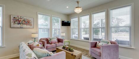 A spacious living room with a large window, a chandelier, and a television mounted on the wall.