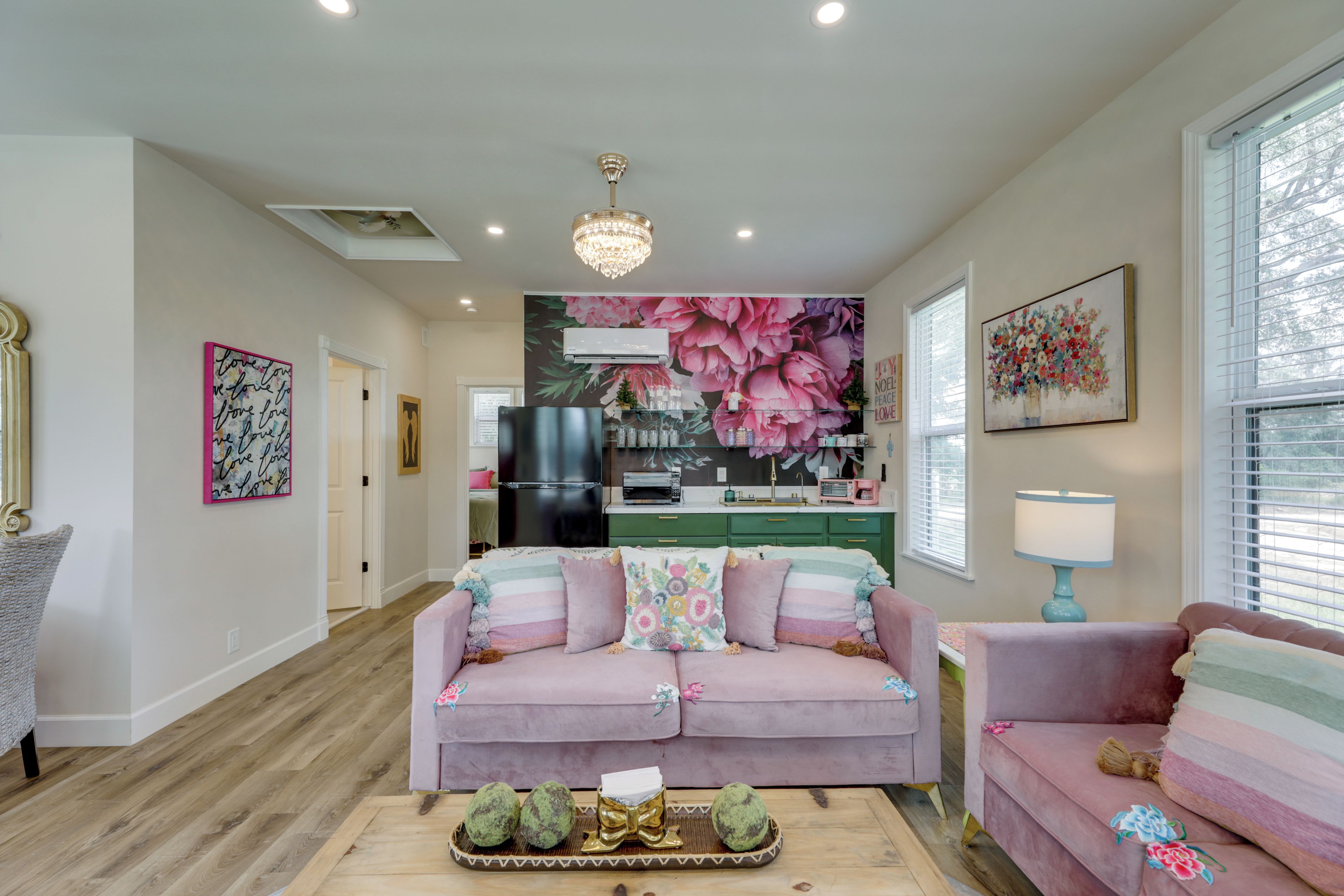 A living room with a pink sofa, a small table with decorative items, a chandelier, and a kitchen area in the background.