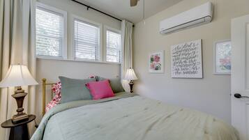 A bedroom with a bed, two bedside lamps, a window with blinds, an air conditioning unit, and a wall with framed artwork.