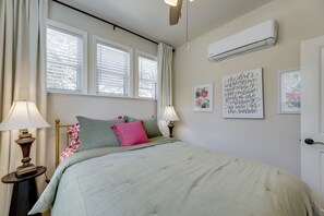 Un dormitorio con cama, dos lámparas de noche, una ventana con persianas, un equipo de aire acondicionado y un cuadro colgado en la pared.