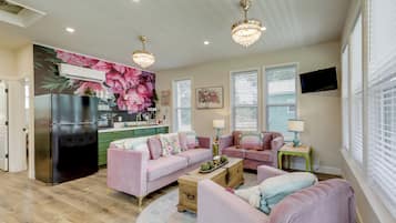 Una sala de estar moderna con un conjunto de sofá rosa, una mesa de centro de madera y una zona de cocina con un refrigerador y un televisor montado en la pared.