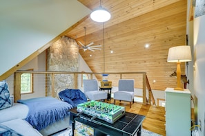 A cozy loft bedroom with a sloped ceiling, a stone wall, a bed with blue bedding, two armchairs, a table with a foosball table, and a lamp.