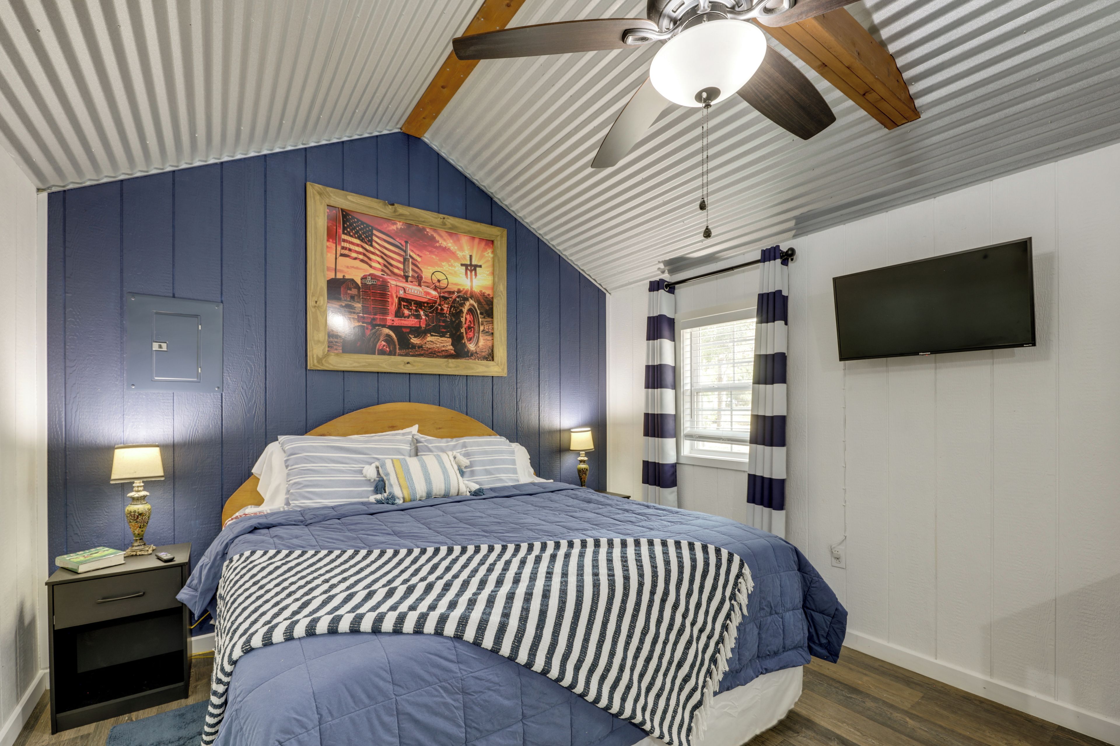 A bedroom with a bed, a ceiling fan, a painting, a TV, and a window with curtains.
