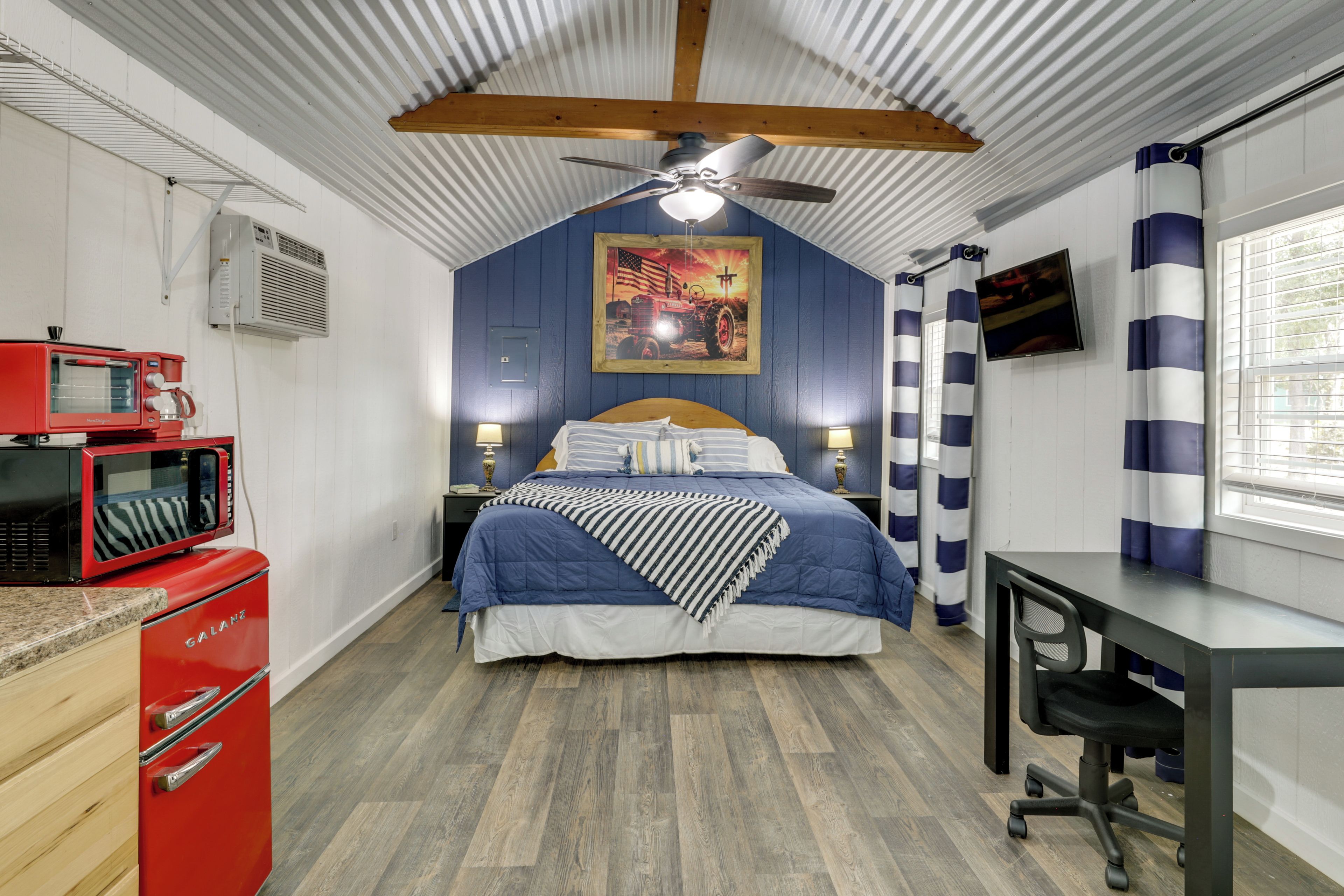 Un dormitorio pequeño con cama, escritorio, microondas rojo y ventilador de techo.