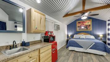 Un dormitorio pequeño con cama, mesitas de noche, un electrodoméstico rojo, ventilador de techo y un ventanal con persianas.