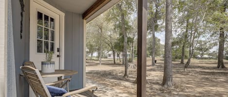 A covered porch with a wooden bench and a table, overlooking a wooded area.