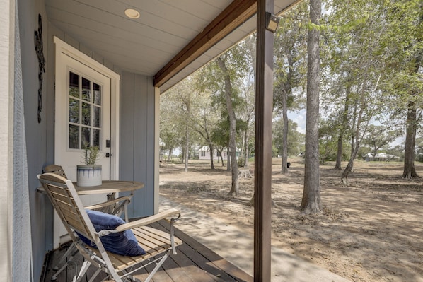 A covered porch with a wooden bench and a table, overlooking a wooded area.