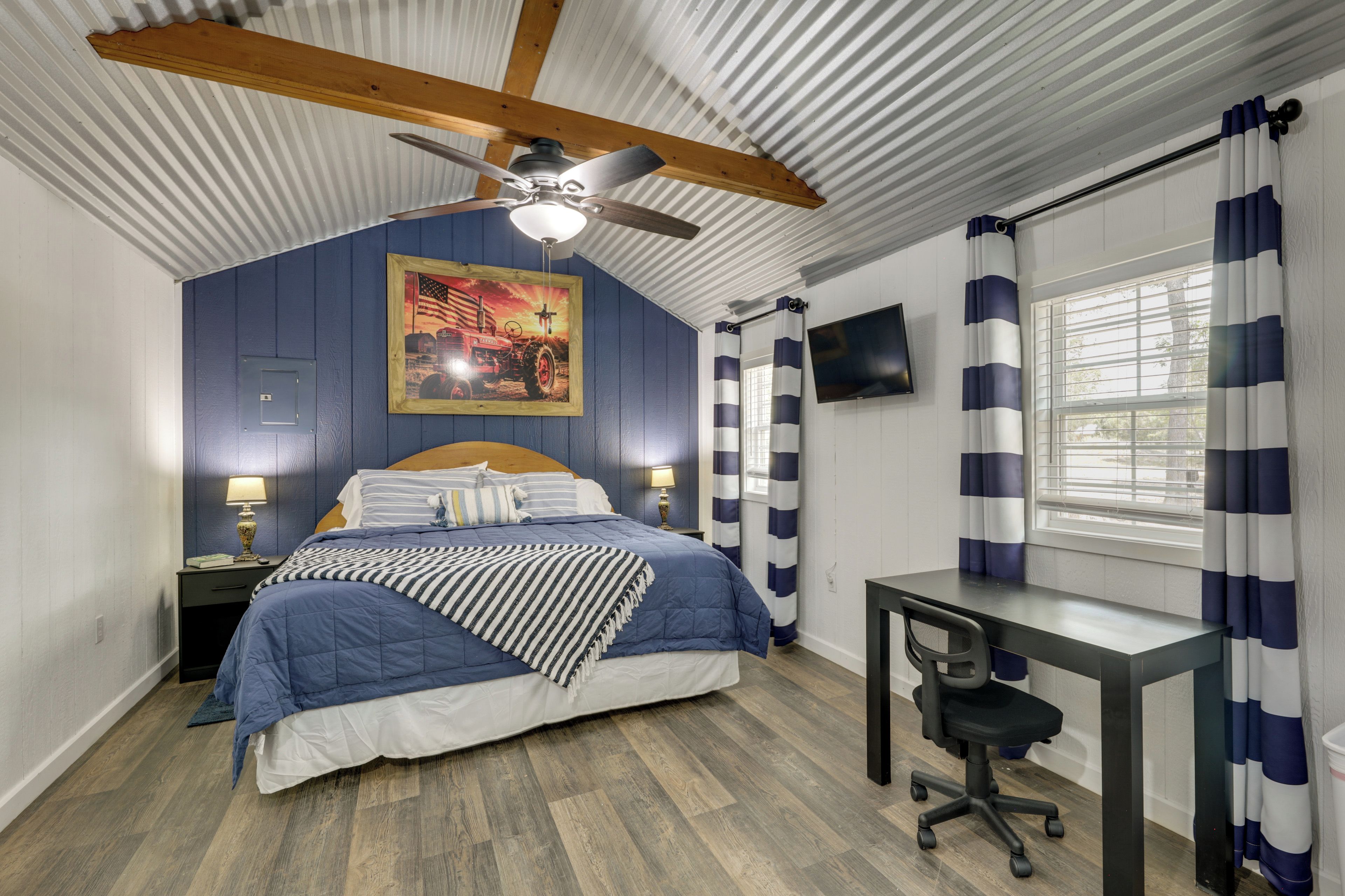 Un dormitorio con cama, escritorio, silla, ventilador de techo, televisor y ventana con cortinas.