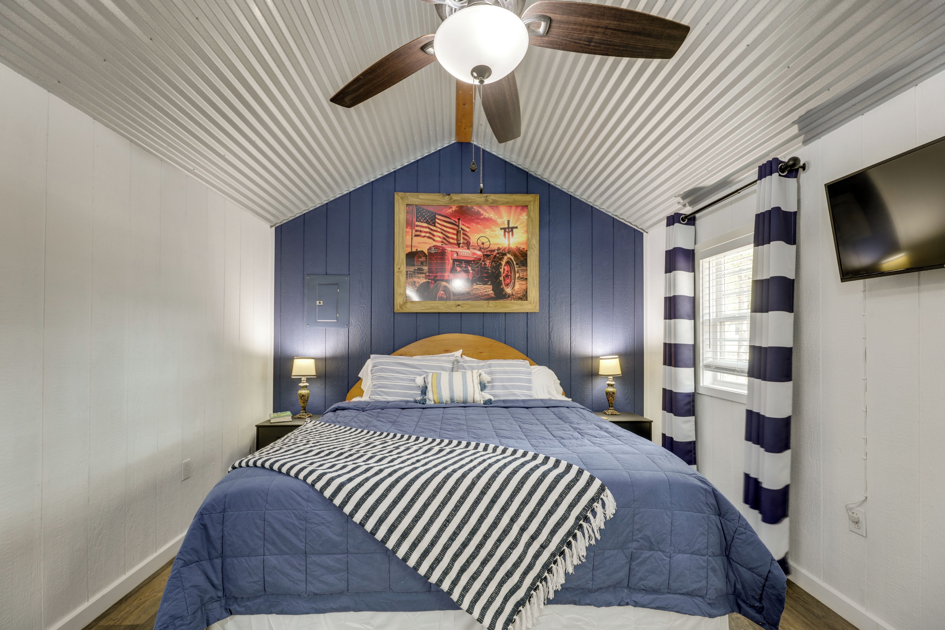 Une chambre avec un lit simple, un ventilateur de plafond, un tableau et une fenêtre avec des rideaux.