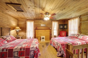 Een kamer in chaletstijl met twee bedden, houten wanden, een plafondventilator, een open haard en een rode stoel.