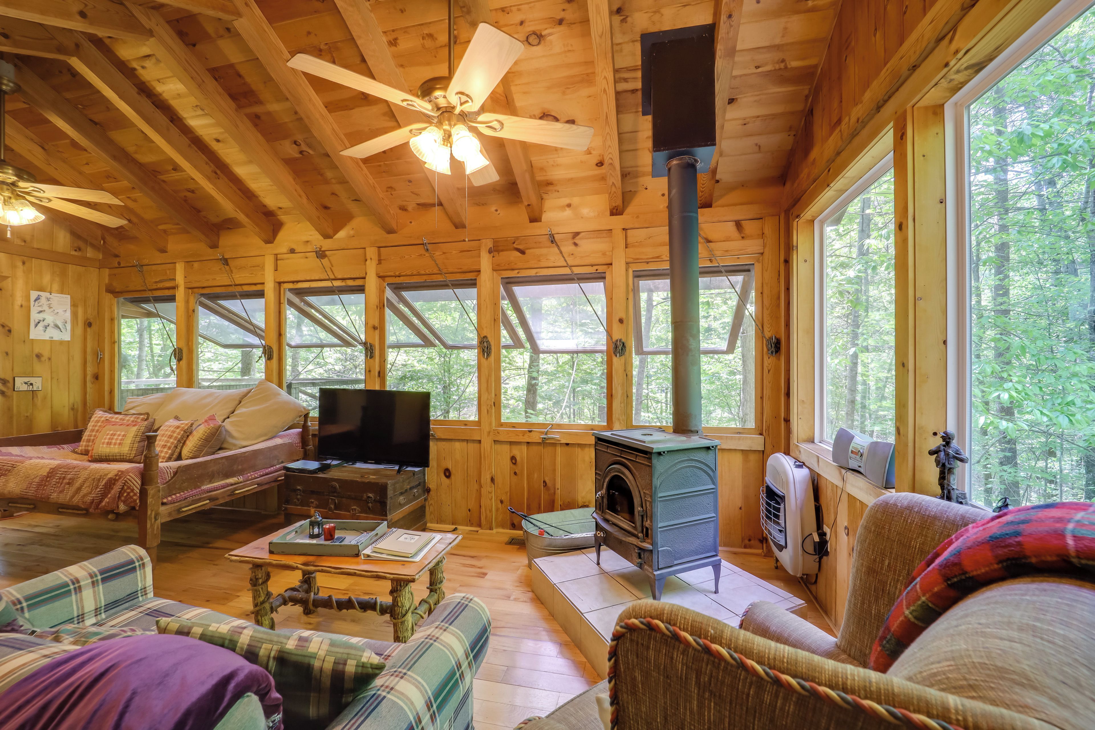 A cozy cabin interior with wooden walls, a ceiling fan, a fireplace, and large windows offering a forest view.