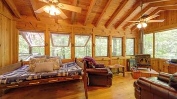 A cozy cabin interior with a wooden floor, a bed with a quilt, a leather sofa, a fireplace, and large windows.