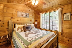 Quarto de cabana de madeira com uma cama coberta por um edredom com tema de urso, uma janela com cortinas e um ventilador de teto.
