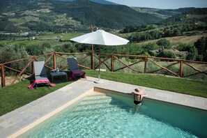 Ein Pool mit Blick auf eine Berglandschaft, eine Holzterrasse mit Liegestühlen und ein Tisch mit einem weißen Sonnenschirm.