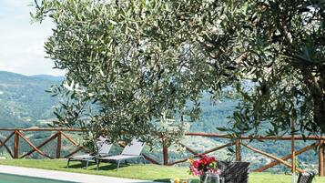 Zona piscina con tavolo e sedie, vista sulle montagne e un albero che offre ombra.