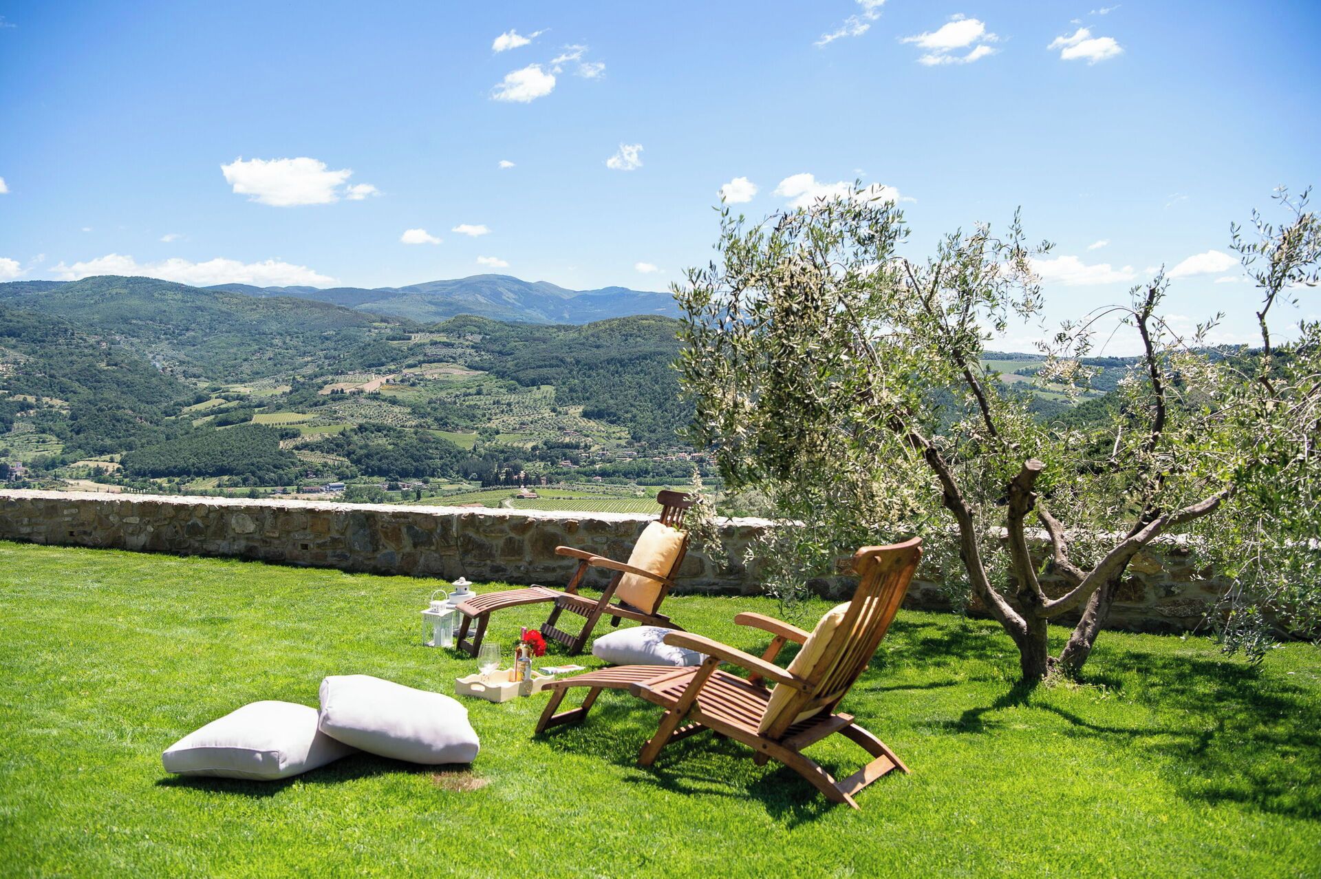 Una zona verde all'aperto con due sedie di legno e un tavolo, con vista su un paesaggio pittoresco con colline e un muro in pietra.