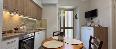 Cocina compacta con comedor, muebles de madera, horno empotrado y un televisor en la pared.