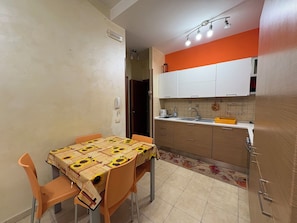Cozinha compacta com armários brancos, pia e bancada. Há uma mesa de jantar com cadeiras e toalha de mesa estampada.