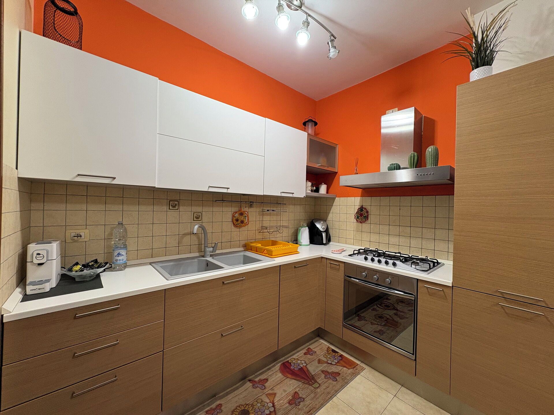 Una cucina con mobili bianchi, un fornello a gas, un lavello e un piano di lavoro con un bollitore e una bottiglia.