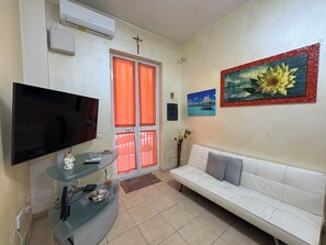 Ein Wohnzimmer mit einem Flachbildfernseher, einem Glastisch, einem weißen Sofa und einer Tür mit orangenen Vorhängen.