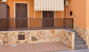 Mural de pedra com grade de metal, escadas que sobem até uma porta e uma janela com contraventanas.