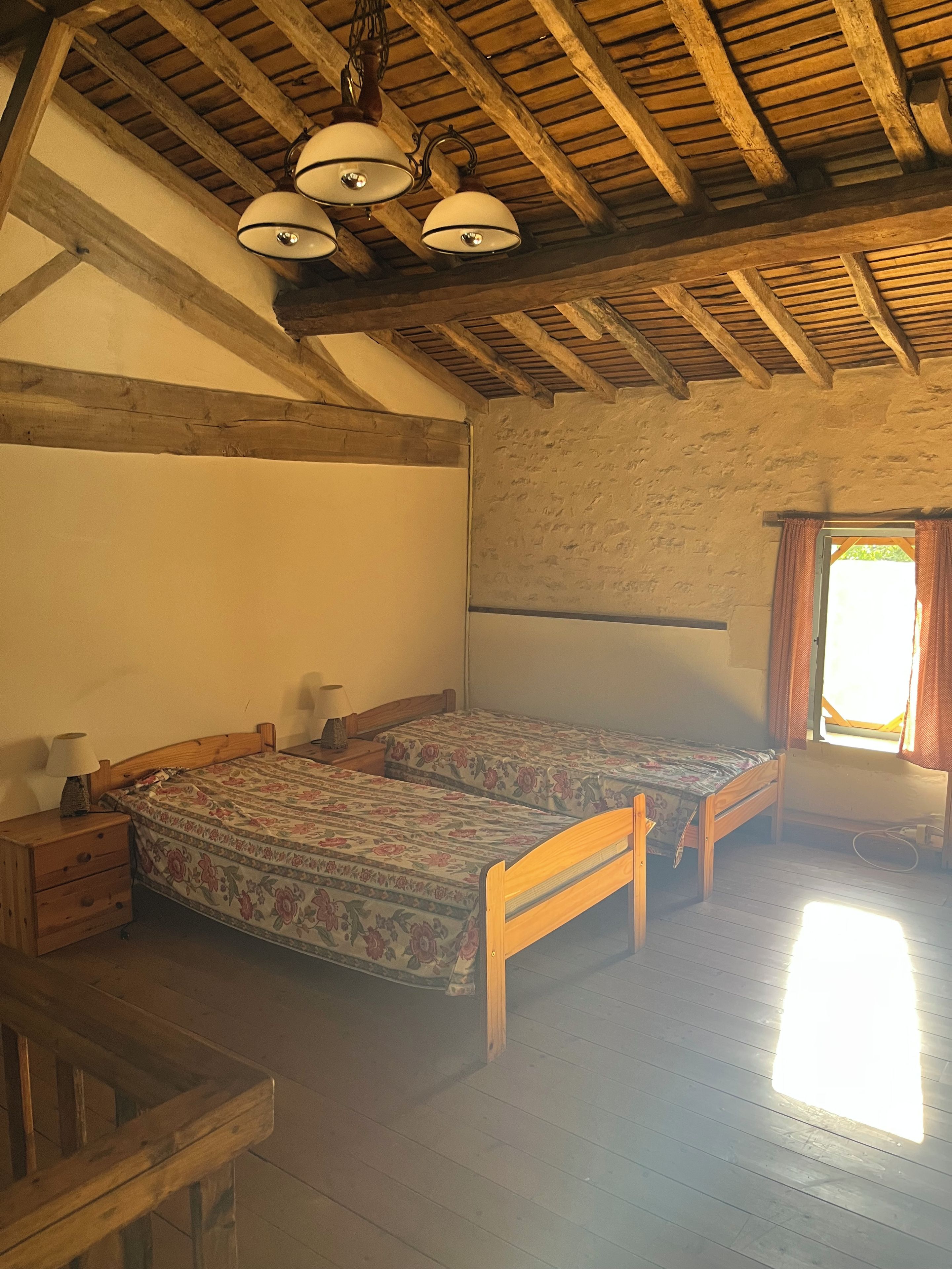 A room with wooden beams, a bed with floral bedding, a nightstand, and a window with curtains.
