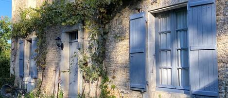 Un bâtiment en pierre doté de nombreuses fenêtres et volets, entouré de verdure et de fleurs.