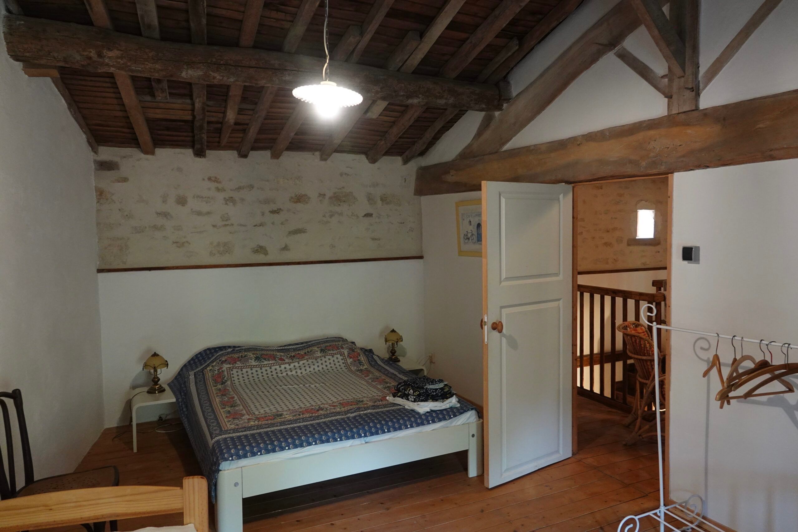 Een slaapkamer met een houten plafond, een bed met een bedovertrek met dessin, een nachtkastje met een lamp en een deur die leidt naar een balkon.