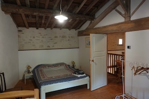 A bedroom with a wooden ceiling, a bed with a patterned bedspread, a nightstand with a lamp, and a door leading to a balcony. - Gite Chez Robin in the Deux-Sèvres (Ardin)