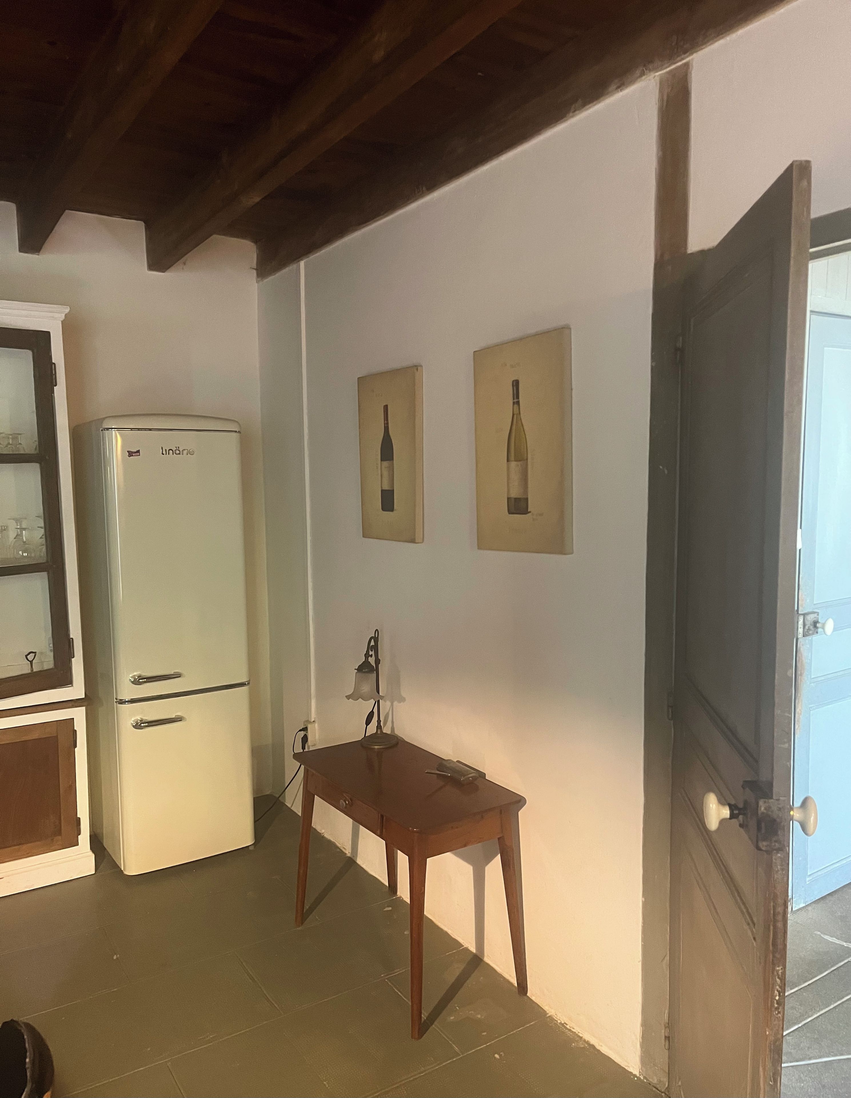 Una habitación con un refrigerador blanco, una mesa de madera con una lámpara, y dos cuadros en la pared.