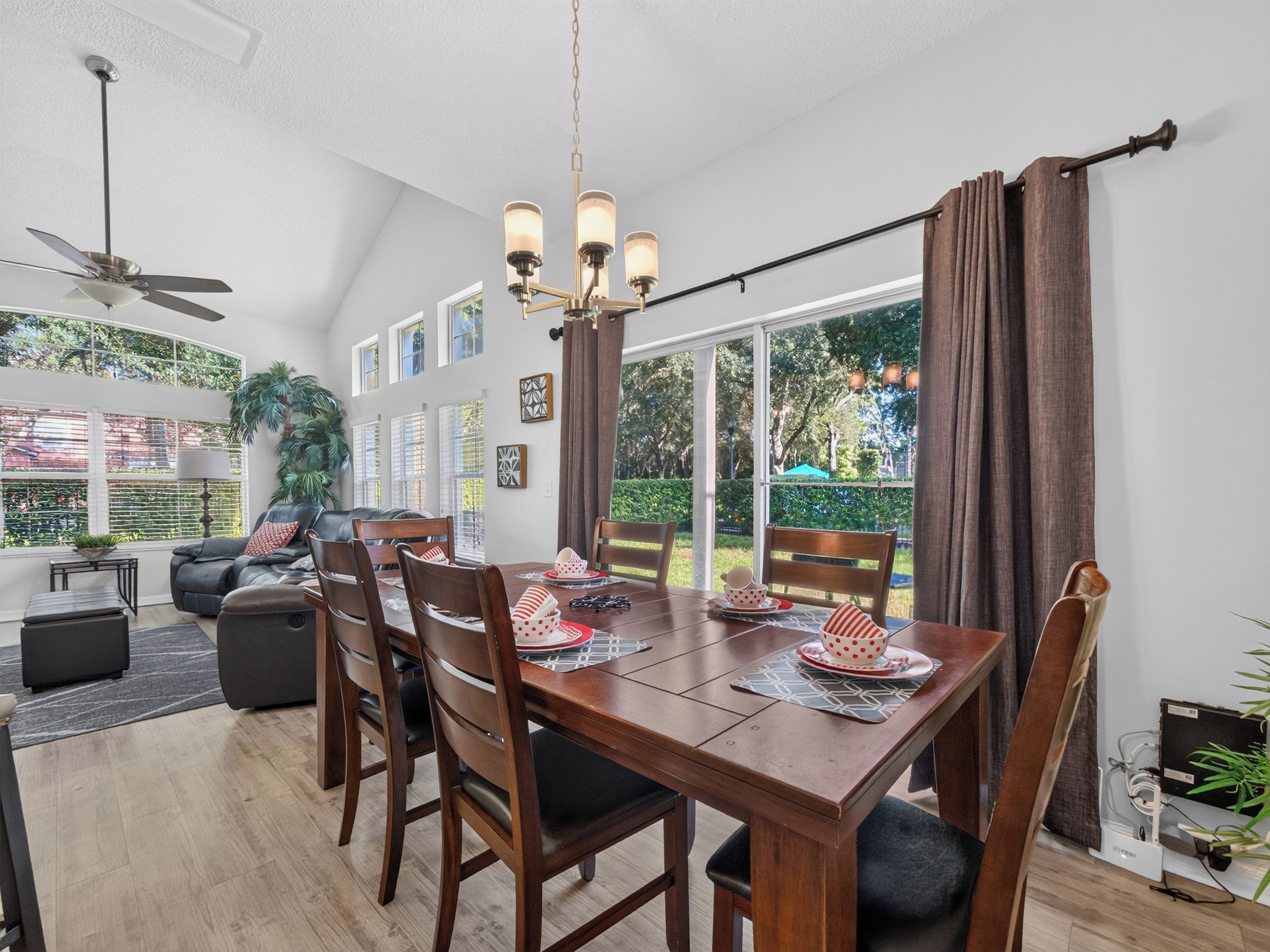 A dining area with a wooden table, chairs, and a chandelier. There is a ceiling fan, a large window with curtains, and a view of greenery outside.