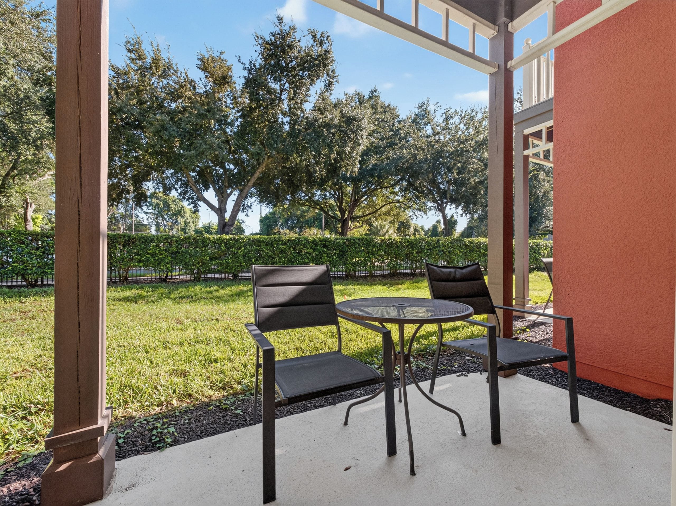 Un patio exterior con dos sillas y una mesa, rodeado de vegetación y un seto.