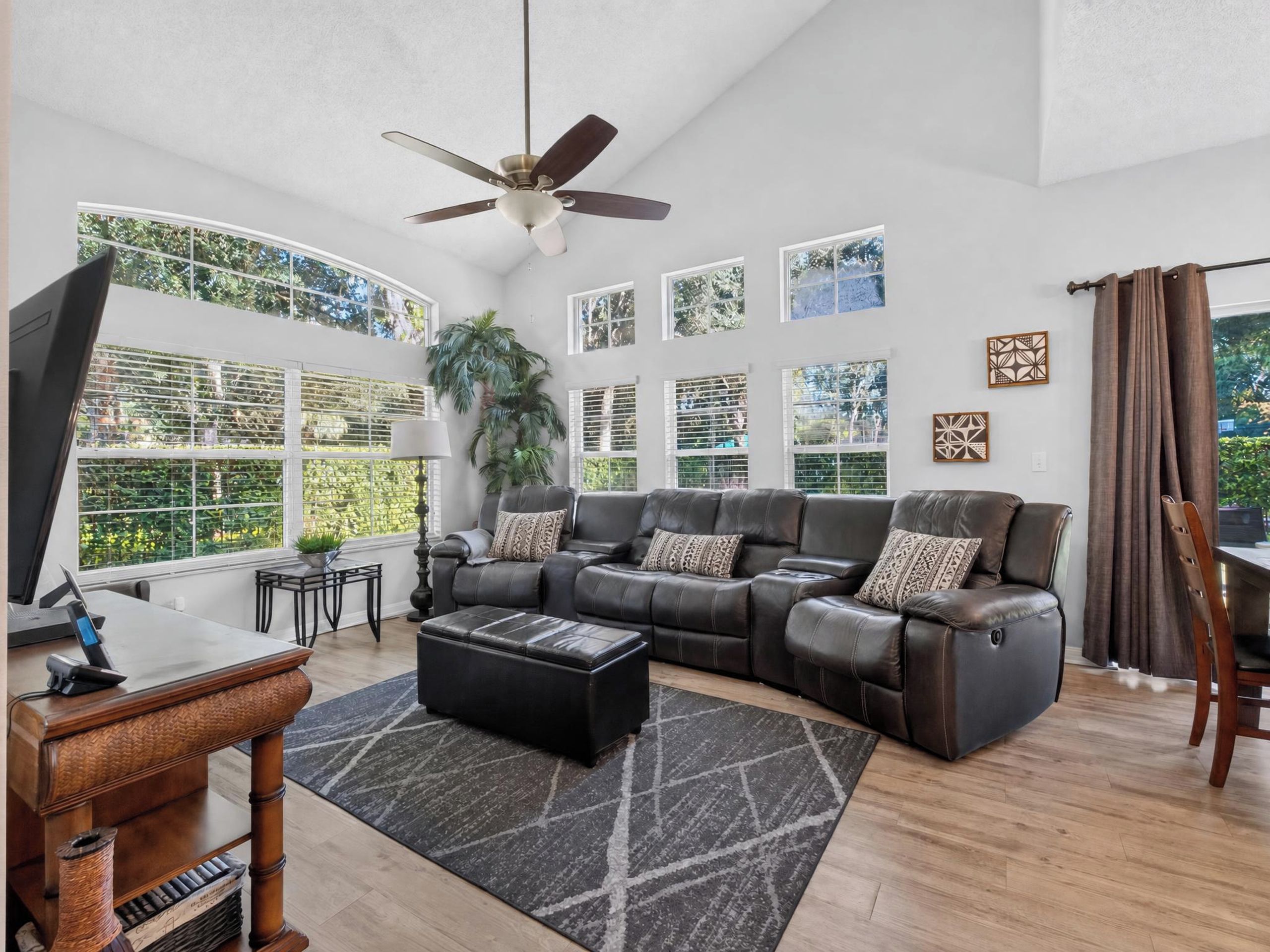 A spacious living room with a large sectional sofa, a ceiling fan, and large windows offering a view of greenery.