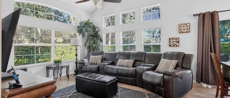 A spacious living room with a large sectional sofa, a ceiling fan, and large windows offering a view of greenery.