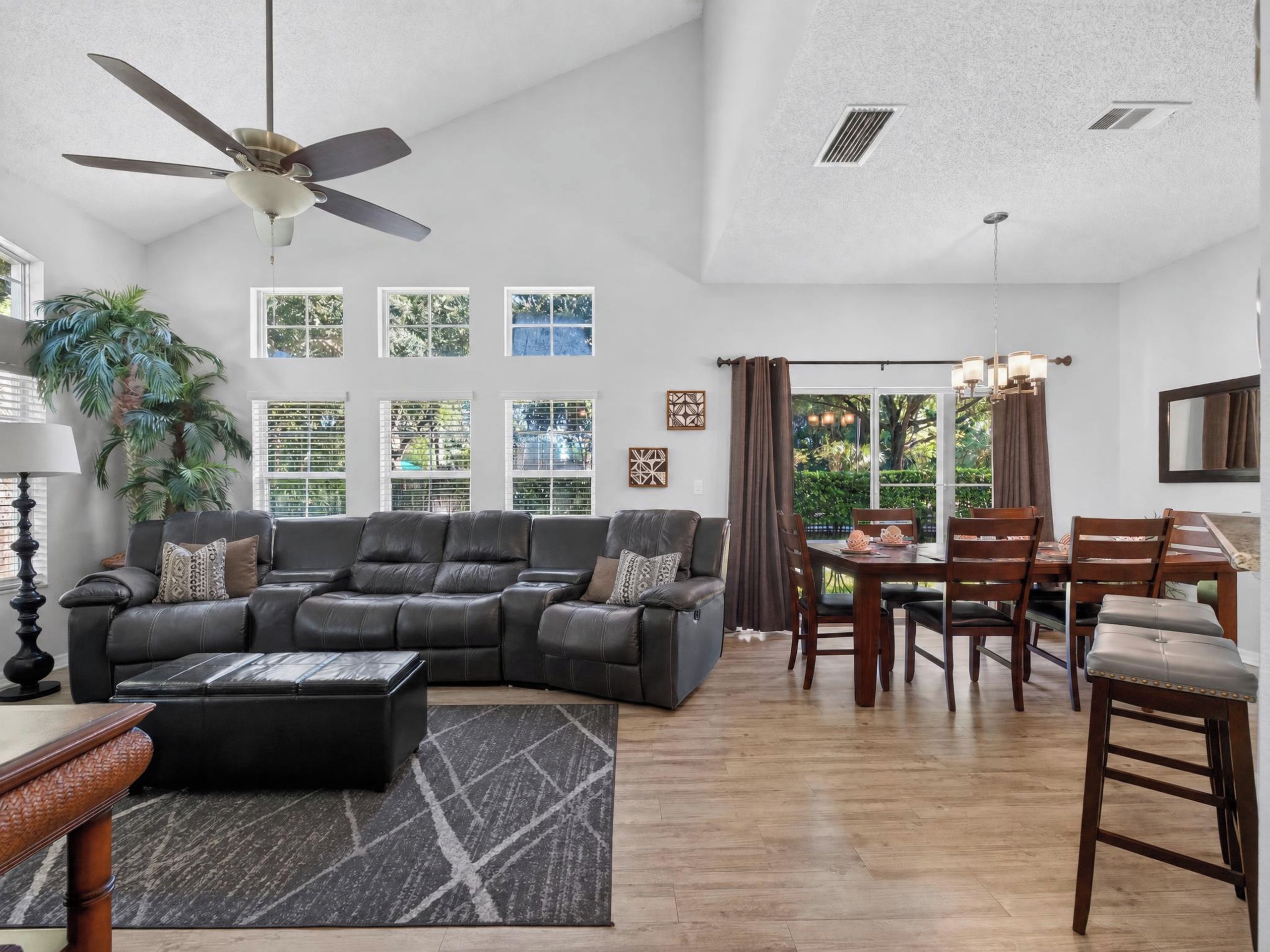 A spacious living room with a sectional sofa, a dining area with wooden chairs, a ceiling fan, and large windows with blinds.