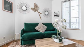 Una habitación con un sofá verde, una mesa de centro de madera, un jarrón blanco con orquídeas y decoración en la pared.