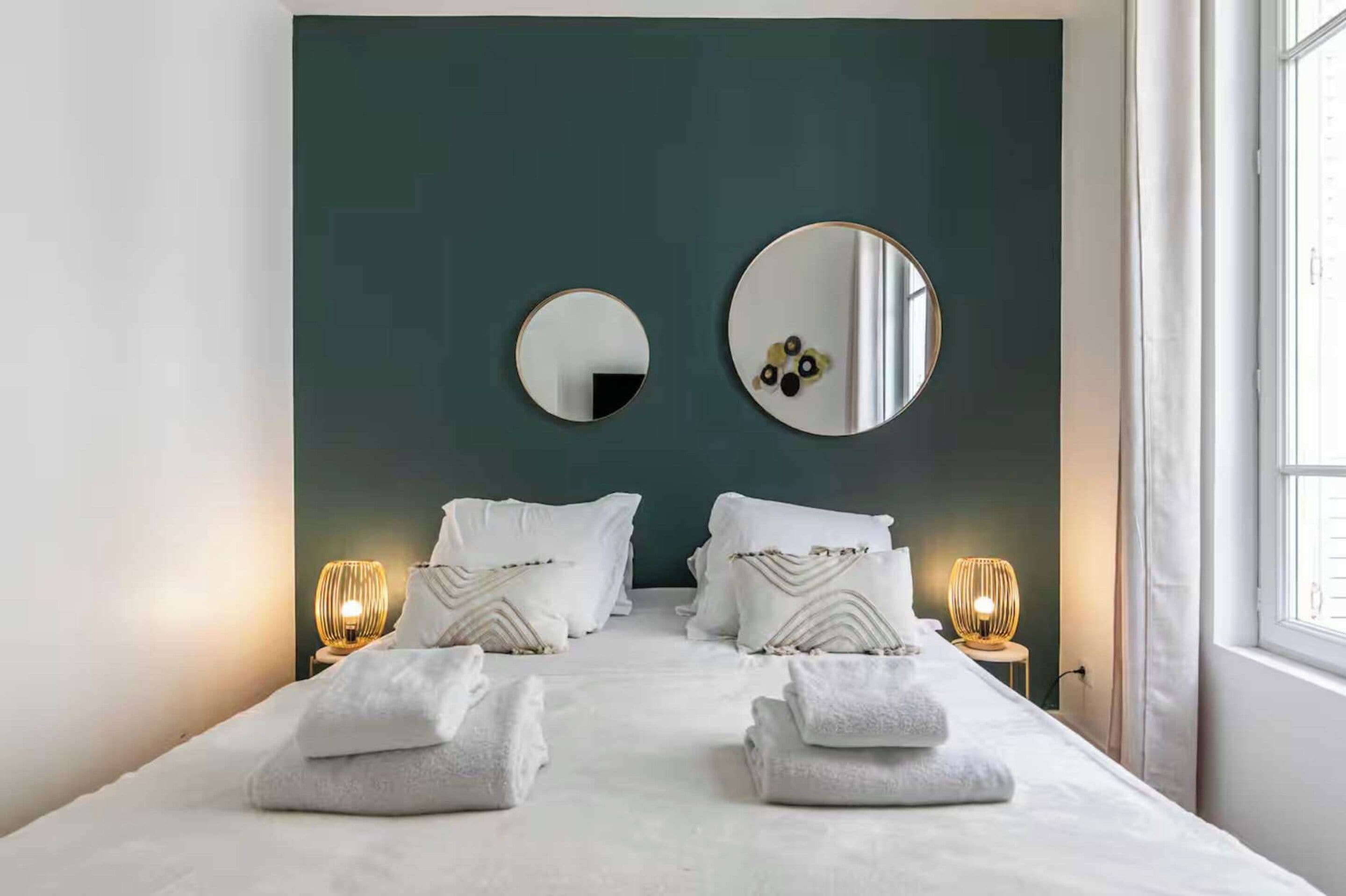 A hotel room with a bed, two bedside lamps, a large mirror, and a window.