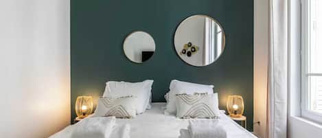 A hotel room with a bed, two bedside lamps, a large mirror, and a window.