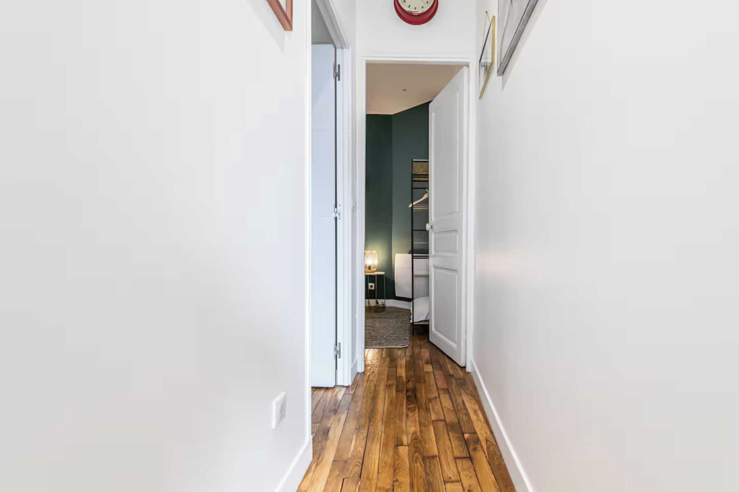 Un pasillo con suelo de madera, paredes blancas y una puerta que da a una habitación con una cama y una mesita de noche.