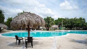 Ein Swimmingpool mit einem Reetdachschirm, Liegestühlen und Palmen.