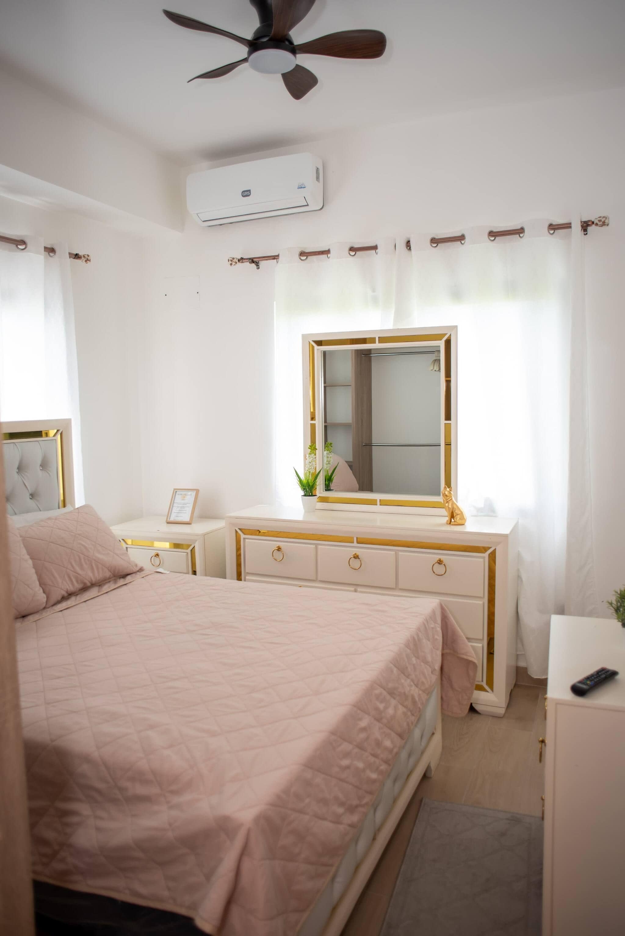 Un dormitorio con cama, tocador con espejo, ventilador de techo, unidad de aire acondicionado y ventana con cortinas.