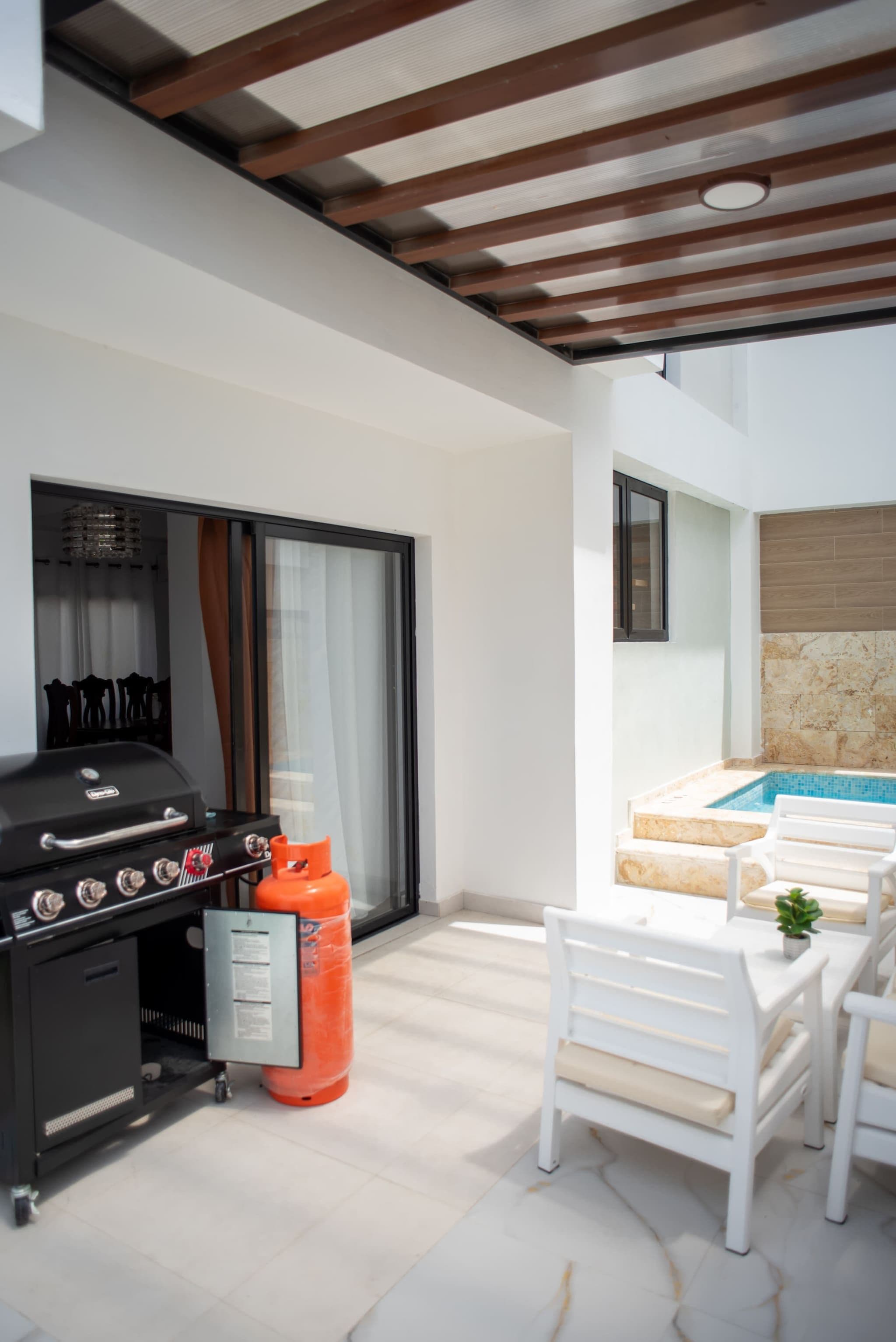 A modern outdoor patio with a barbecue grill, a gas cylinder, white outdoor furniture, and a swimming pool.