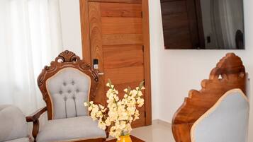A living room with a sofa, two armchairs, a coffee table, a vase with flowers, a wooden door, and a television mounted on the wall.