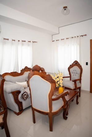 Una sala de estar con sofá, sillón y mesa de centro, un jarrón con flores y cortinas.