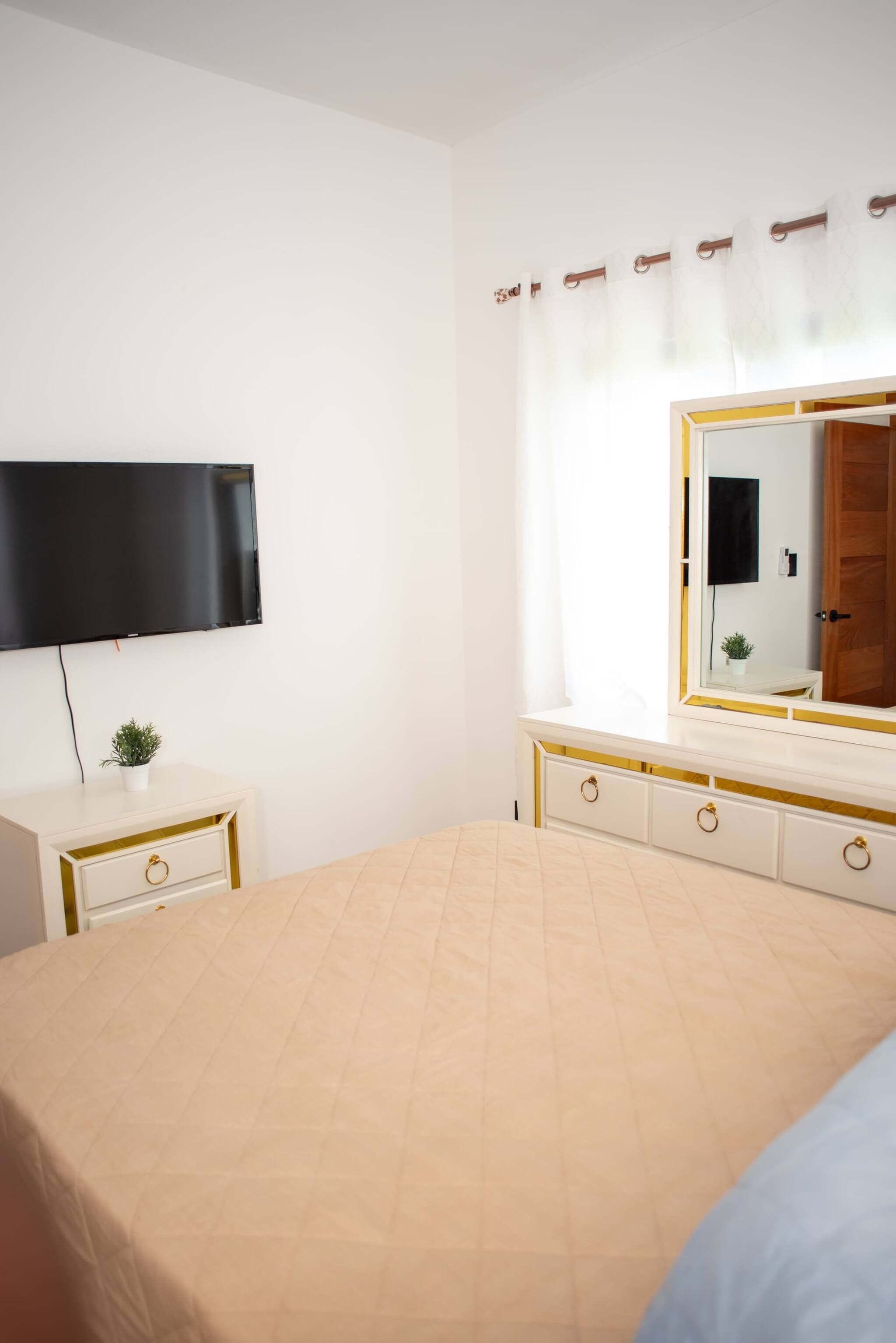 Une chambre à coucher avec un lit, un téléviseur fixé au mur, une table de chevet avec une plante et un grand miroir avec un cadre doré.